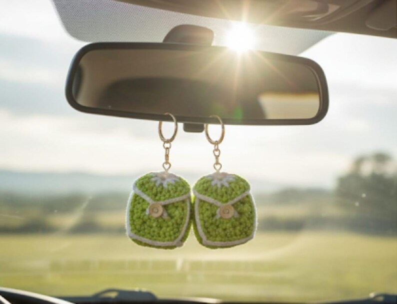 Op de afbeelding: Twee groen-witte gehaakte auto-hangers hangen aan een achteruitkijkspiegel. De hangers zijn vierkant met knoopdetails en een witte rand. De achtergrond toont een zonnige buitensc&egrave;ne.
