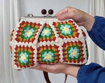 Puede incluir: Bolso de crochet hecho a mano con cierre metálico y cuentas de madera marrón. El bolso presenta un diseño de cuadrados de la abuela coloridos con cuadrados rojos, verdes, amarillos y azules. Tiene un fondo crema y un asa de cuero marrón.