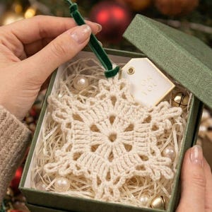 Op de afbeelding: Een crèmekleurig gehaakt sneeuwvlokornament met een donkergroene fluwelen lintlus, geplaatst in een vierkante groene geschenkdoos gevuld met versnipperd papier, kleine witte parels en een cadeaulabel met de tekst FOR YOU erop gedrukt.