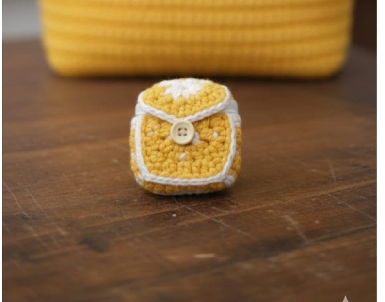 Op de afbeelding: Een kleine, handgemaakte gehaakte tas in geel en wit. De tas heeft een knoopsluiting en een witte rand. Op de achtergrond een groter gehaakt geel item, mogelijk een mand, op een houten oppervlak.