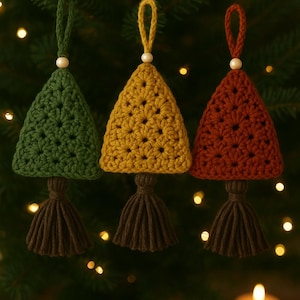 Op de afbeelding: Drie handgemaakte gehaakte kerstboomversieringen in groen, geel en rood-oranje, elk met een bijpassende kwast en een houten kraal. De ornamenten hangen aan een kerstboom met zachte, vage lichten op de achtergrond.