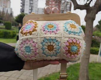 Puede incluir: Un bolso de ganchillo con cuadrados de abuela, marco de madera y una correa larga. El bolso es blanco con detalles en rosa, azul y marrón.