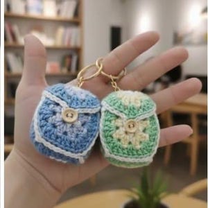 Op de afbeelding: Twee kleine, gehaakte rugzak-sleutelhangers. E&eacute;n is blauw en wit, de andere groen en cr&egrave;me. Beide hebben een knoopsluiting en goudkleurige sleutelringen. De sleutelhangers worden in een open hand gehouden.