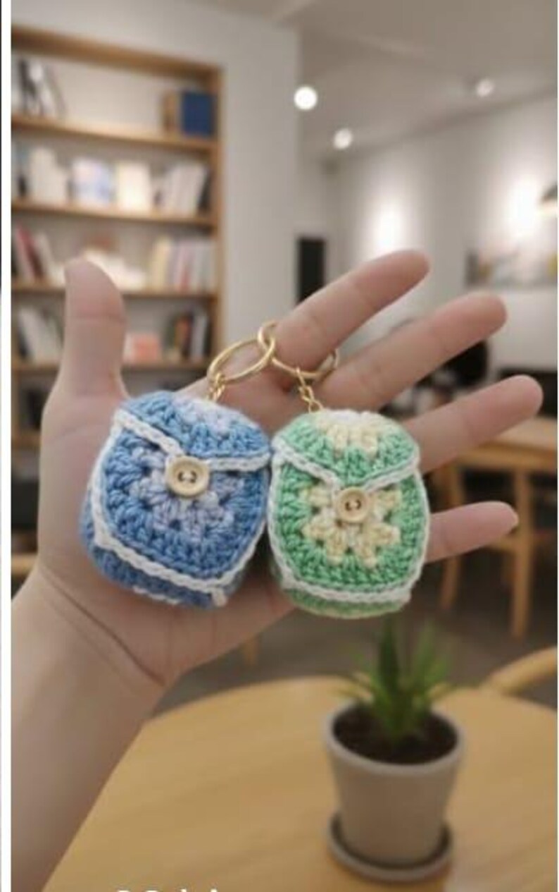 Op de afbeelding: Twee kleine, gehaakte rugzak-sleutelhangers. E&eacute;n is blauw en wit, de andere groen en cr&egrave;me. Beide hebben een knoopsluiting en goudkleurige sleutelringen. De sleutelhangers worden in een open hand gehouden.