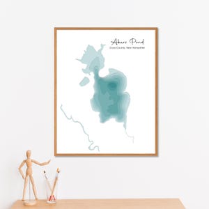 Puede incluir: Una impresión enmarcada de Akers Pond en el condado de Coos, New Hampshire. El mapa es de color azul verdoso y muestra las curvas de nivel de profundidad del estanque. La impresión está en un marco de madera y se muestra en una pared blanca.