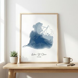 May include: A framed print showing a blue watercolour map of Lake St. Clair, Canada. The artwork is in a light wood frame against a white backdrop. The text "Lake St. Clair" and "Canada" are printed below the map.