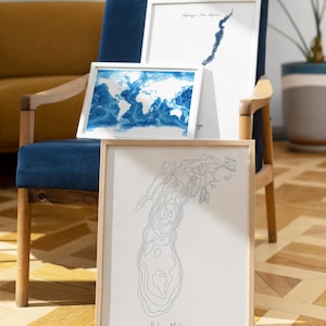 May include: Framed art prints featuring a world map, a lake contour map, and a coastal outline. The prints are in white frames, with blue and gray tones. The Lake Michigan print includes the text "Lake Michigan, United States."