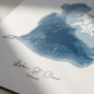 May include: A watercolour print of Lake St. Clair in shades of blue. The lake's contours are depicted with layered detail, creating a topographical effect. The text "Lake St. Clair, Canada" is written in an elegant script at the bottom of the print.