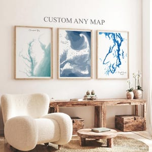 May include: Three framed coastal map prints in shades of blue and green hang on a white wall. The prints are labeled "Chesapeake Bay", "Cape Cod", and "Puget Sound". The text "CUSTOM ANY MAP" is above the prints. A cream-colored chair and wooden furniture are in the foreground.