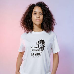May include: White t-shirt featuring a black graphic of a crown of thorns and the words "EL CAMINO LA VERDAD LA VIDA" with "Juan 14:6" below. The shirt is worn by a person with curly dark hair.