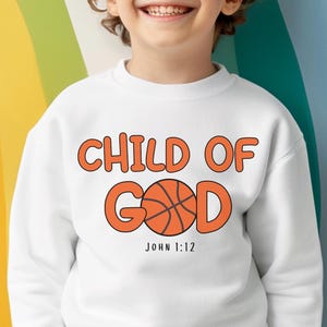 Puede incluir: Sudadera blanca con la frase "CHILD OF GOD" en naranja, con un gráfico de baloncesto. El texto "JOHN 1:12" está debajo. La imagen es un archivo digital. La sudadera la lleva un niño.