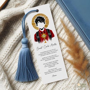 Pode incluir: Um marcador de livros branco com uma borla azul. O marcador de livros apresenta uma ilustração de São Carlo Acutis com o texto "Saint Carlo Acutis" e uma oração. A borla é feita de fio azul.