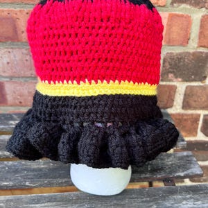 May include: A handmade crochet hat with a black top, red middle, yellow band, and a black ruffled brim. The hat is displayed on a white, rounded object, likely a mannequin head, against a brick wall background.
