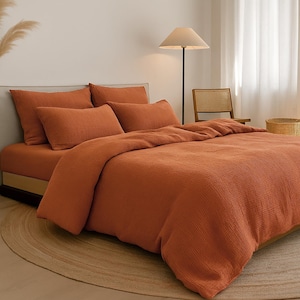 以下が含まれることがあります： テラコッタ色の寝具がセットされたベッド。掛け布団カバーと枕が含まれています。ベッドは丸いベージュのラグの上にあります。フロアランプと籐の椅子が背景にあります。部屋はニュートラルなカラーパレットです。