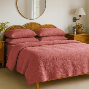 Op de afbeelding: Een bed met een rood-oranje gewatteerde sprei en bijpassende kussens. Het bed heeft een houten frame en hoofdbord. Een ronde spiegel hangt aan de muur boven het bed. Een houten nachtkastje met een lamp en bloemen staat naast het bed.