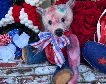 May include: A handcrafted teddy bear in shades of pink, blue, and red, sporting a red and blue checked bow tie and blue denim trousers. The bear has a key pendant and sits before a brick wall, surrounded by patriotic decor.