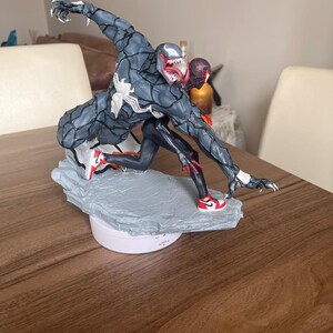 May include: A detailed figurine of Venom and Spider-Man in action. Venom is mainly grey with a white spider symbol, and Spider-Man is in a black and red suit. Both figures are wearing red and white trainers. The base is a light grey colour.