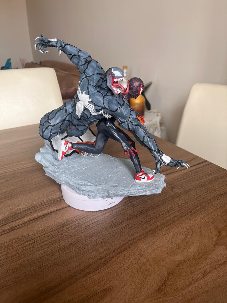 May include: A detailed figurine of Venom and Spider-Man in action. Venom is mainly grey with a white spider symbol, and Spider-Man is in a black and red suit. Both figures are wearing red and white trainers. The base is a light grey colour.
