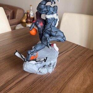 May include: A dynamic figurine featuring two comic book characters in action. One figure is a dark blue and grey Venom, and the other is a red and black Spider-Man holding an orange basketball. The base is a grey, rocky structure.