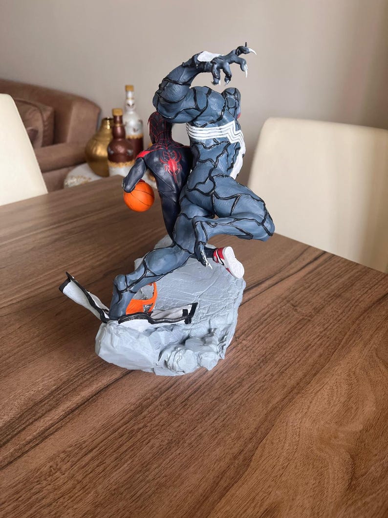 May include: A dynamic figurine featuring two comic book characters in action. One figure is a dark blue and grey Venom, and the other is a red and black Spider-Man holding an orange basketball. The base is a grey, rocky structure.