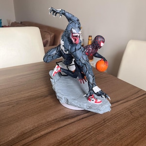 May include: A detailed figurine of Venom and Spider-Man playing basketball. Venom, in a dark grey and black colour scheme, is leaping with a basketball. Spider-Man is in a red and black suit, also holding a basketball. Both are wearing trainers.