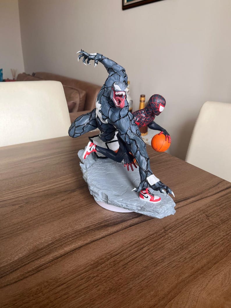 May include: A detailed figurine of Venom and Spider-Man playing basketball. Venom, in a dark grey and black colour scheme, is leaping with a basketball. Spider-Man is in a red and black suit, also holding a basketball. Both are wearing trainers.