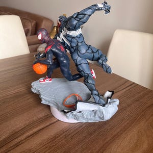May include: A detailed figurine depicting two characters in action. One character is in a black and red suit, dribbling an orange basketball. The other character is a dark grey figure with white accents. The base is grey and white, with a small basketball hoop.