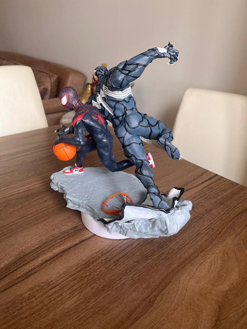 May include: A detailed figurine depicting two characters in action. One character is in a black and red suit, dribbling an orange basketball. The other character is a dark grey figure with white accents. The base is grey and white, with a small basketball hoop.