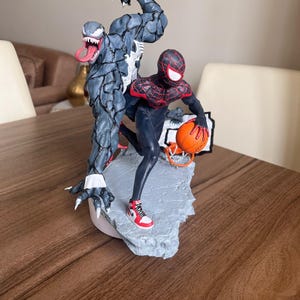 May include: A detailed figurine featuring Spider-Man and Venom in a dynamic pose. Spider-Man is holding a basketball, while Venom looms behind. The statue is painted in shades of black, red, and grey, with intricate detailing.