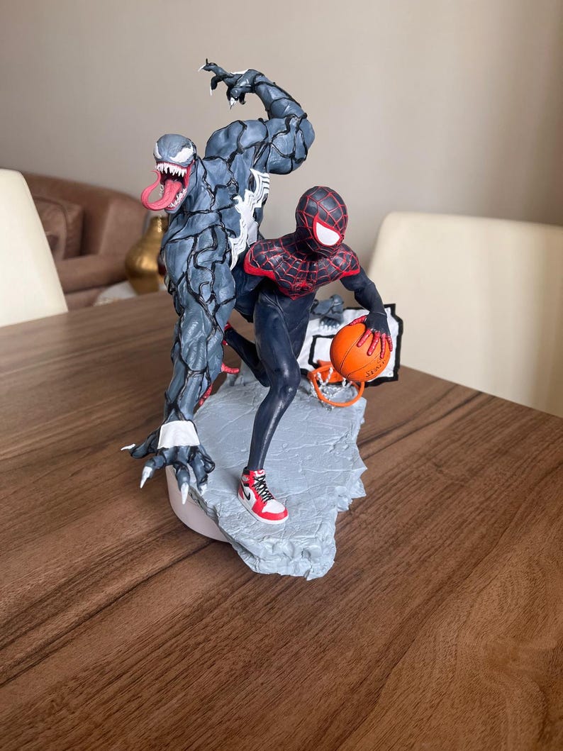 May include: A detailed figurine featuring Spider-Man and Venom in a dynamic pose. Spider-Man is holding a basketball, while Venom looms behind. The statue is painted in shades of black, red, and grey, with intricate detailing.