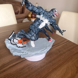 May include: A detailed figurine of Venom, a Marvel Comics character, in a dynamic pose. The figure is primarily dark blue with white accents and a long red tongue. It stands on a textured, light blue base, with a figure of Spider-Man. The base is on a white circular platform.