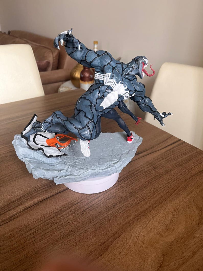 May include: A detailed figurine of Venom, a Marvel Comics character, in a dynamic pose. The figure is primarily dark blue with white accents and a long red tongue. It stands on a textured, light blue base, with a figure of Spider-Man. The base is on a white circular platform.