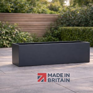 May include: A dark grey rectangular planter box, made in Britain. The planter is placed on a stone patio, with a wooden fence and green foliage in the background. The planter is a simple, modern design.