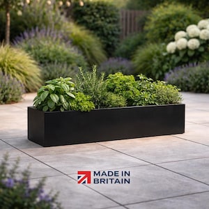 May include: A black rectangular planter box filled with a variety of green herbs. The planter is made of a dark material and sits on a tiled patio. The background features greenery and the text "MADE IN BRITAIN".