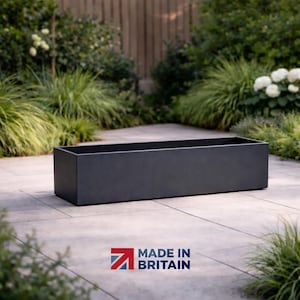 May include: A dark grey rectangular planter box, with a simple, modern design. The planter is made of a solid material and is positioned on a stone patio, surrounded by plants. The text "MADE IN BRITAIN" is visible.