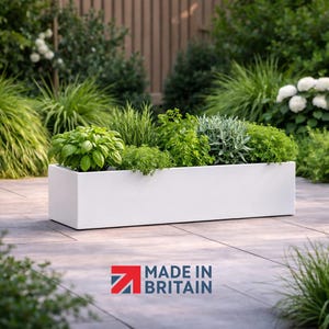 May include: A white rectangular planter box filled with a variety of green plants. The box sits on a stone patio, surrounded by greenery. The text "MADE IN BRITAIN" is visible in the lower right corner.