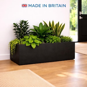 May include: A black rectangular planter box filled with a variety of green plants. The planter is on a wooden floor near a doorway. The text "MADE IN BRITAIN" is displayed above the planter. The planter is filled with plants of different shapes and sizes.