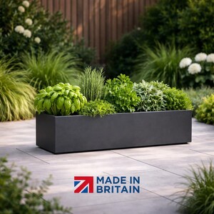 May include: A dark grey rectangular planter box overflowing with a variety of green herbs and plants. The planter sits on a patio, with a backdrop of green foliage. The text "MADE IN BRITAIN" is visible in the bottom right corner.