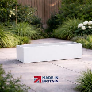 May include: A white rectangular planter box, with a minimalist design. The planter is made of a solid material and is set on a paved patio, surrounded by plants. The text "MADE IN BRITAIN" is visible. The planter is suitable for outdoor use.