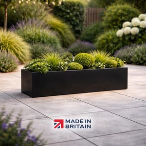May include: A black rectangular planter box filled with a variety of green plants. The box is positioned on a tiled patio, with a backdrop of garden foliage. The text "MADE IN BRITAIN" is displayed in the lower right corner of the image.