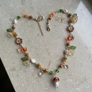 Könnte beinhalten: Eine goldfarbene Kettenhalskette mit einem zentralen Pilzanhänger, durchsetzt mit verschiedenen Perlen, darunter grüne Blätter, durchscheinende orangefarbene und klare Blumen, braune Holzkugeln und kleine weiße tropfenförmige Perlen.