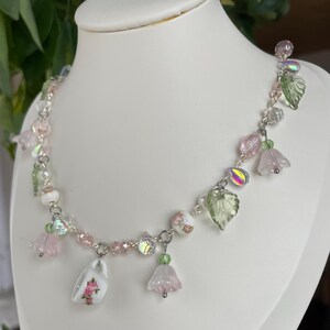 Op de afbeelding: Een delicate ketting met een zilveren ketting, roze en heldere kralen, iriserende accenten en bloemenbedels. De ketting bevat kleine roze bloemhangers, groene bladvormige bedels en een miniatuur theekopjes hanger met een bloemenontwerp.