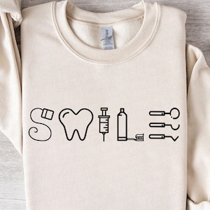 May include: A cream-colored sweatshirt with a black graphic of dental tools and the word "SMILE".