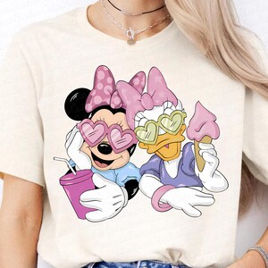 May include: Cream-colored t-shirt featuring Minnie Mouse and Daisy Duck. Minnie wears heart-shaped sunglasses and holds a drink. Daisy Duck wears heart-shaped sunglasses and holds an ice cream cone. Both characters are wearing bows.