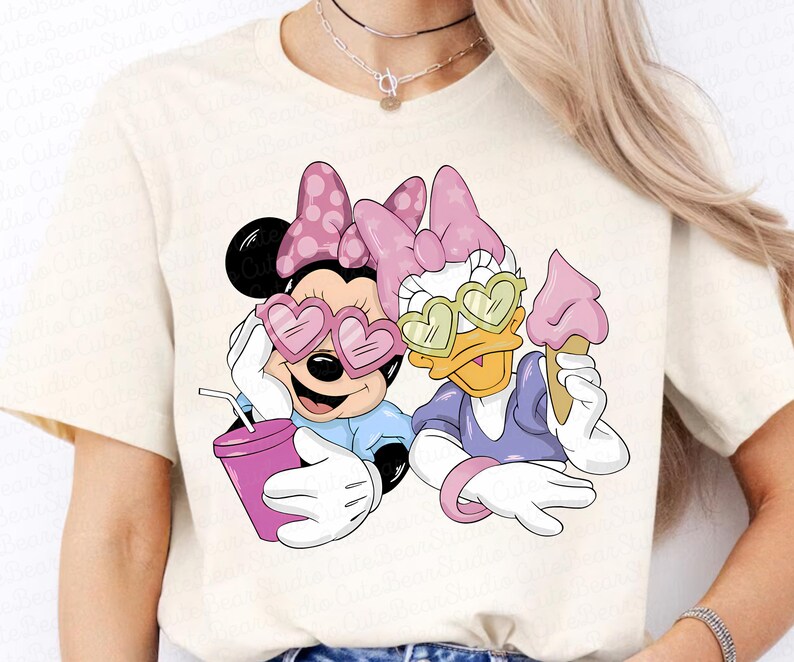May include: Cream-colored t-shirt featuring Minnie Mouse and Daisy Duck. Minnie wears heart-shaped sunglasses and holds a drink. Daisy Duck wears heart-shaped sunglasses and holds an ice cream cone. Both characters are wearing bows.
