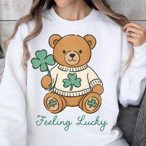 May include: White sweatshirt featuring a cartoon teddy bear holding a green shamrock. The bear wears a sweater with a shamrock design. The words "Feeling Lucky" are written in green script below the bear.