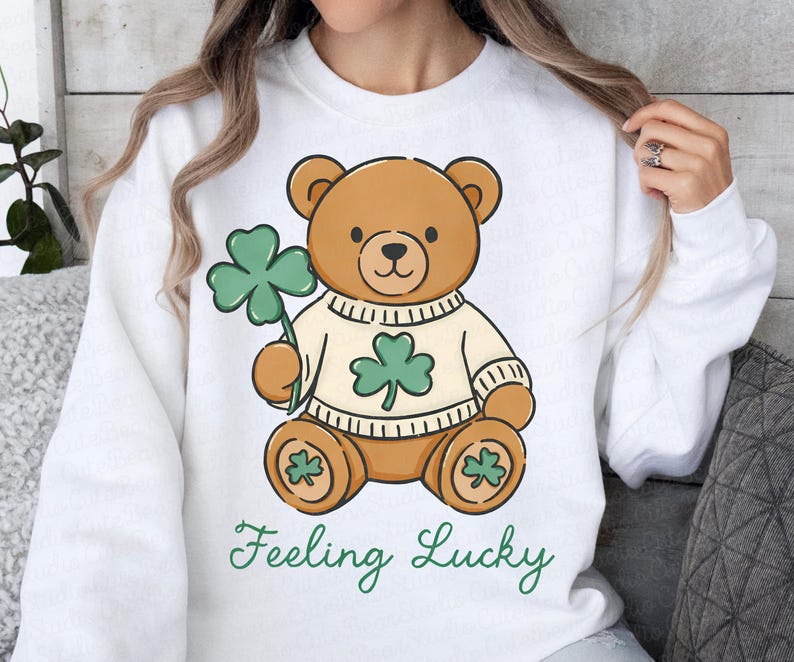May include: White sweatshirt featuring a cartoon teddy bear holding a green shamrock. The bear wears a sweater with a shamrock design. The words "Feeling Lucky" are written in green script below the bear.