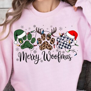 May include: A light pink sweatshirt with a festive design. The graphic features paw prints decorated with Christmas lights, Santa hats, and reindeer antlers. The words "Merry Woofmas" are written in a script font.