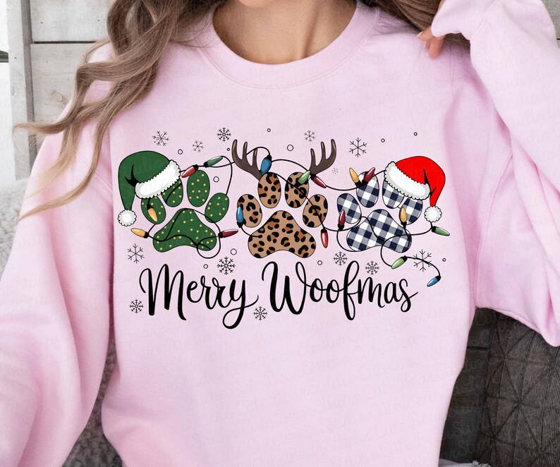 May include: A light pink sweatshirt with a festive design. The graphic features paw prints decorated with Christmas lights, Santa hats, and reindeer antlers. The words "Merry Woofmas" are written in a script font.