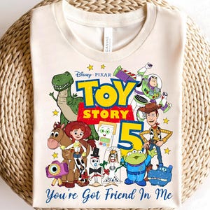 May include: A cream-colored t-shirt featuring a colorful graphic of Disney Pixar Toy Story characters including Woody, Buzz Lightyear, Jessie, Rex, Bullseye, and others. The design includes the text Toy Story 5 and You've Got Friend In Me.